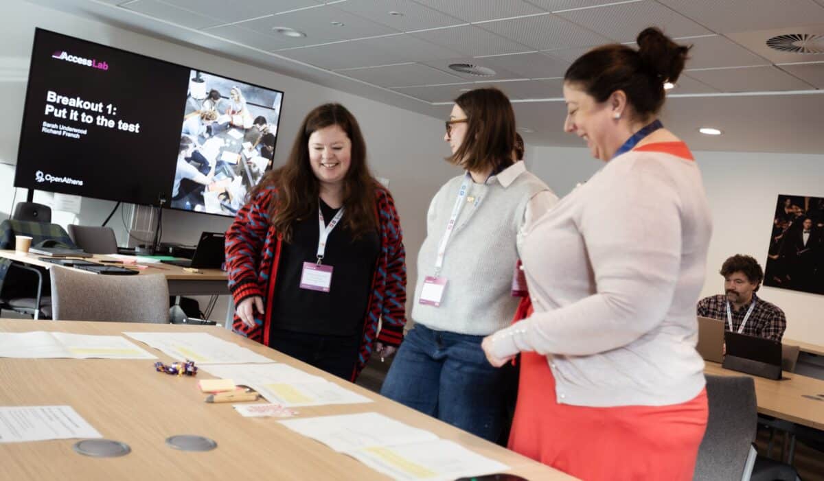 3 women standing around a table, preparing for the breakout room session at Access Lab EMEA.
